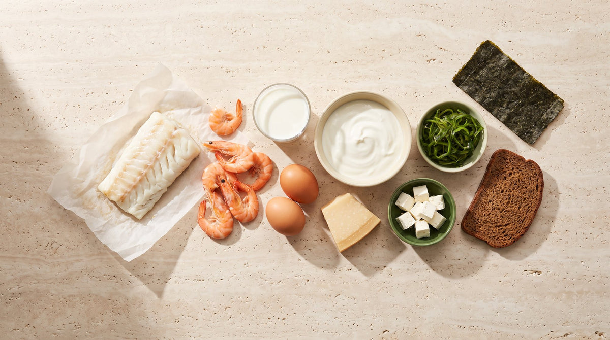 Jodreiche Lebensmittel auf hellem Stein: Kabeljau, Garnelen, Eier, Milch, Joghurt, Parmesan, Feta, Roggenbrot und ein Nori-Blatt