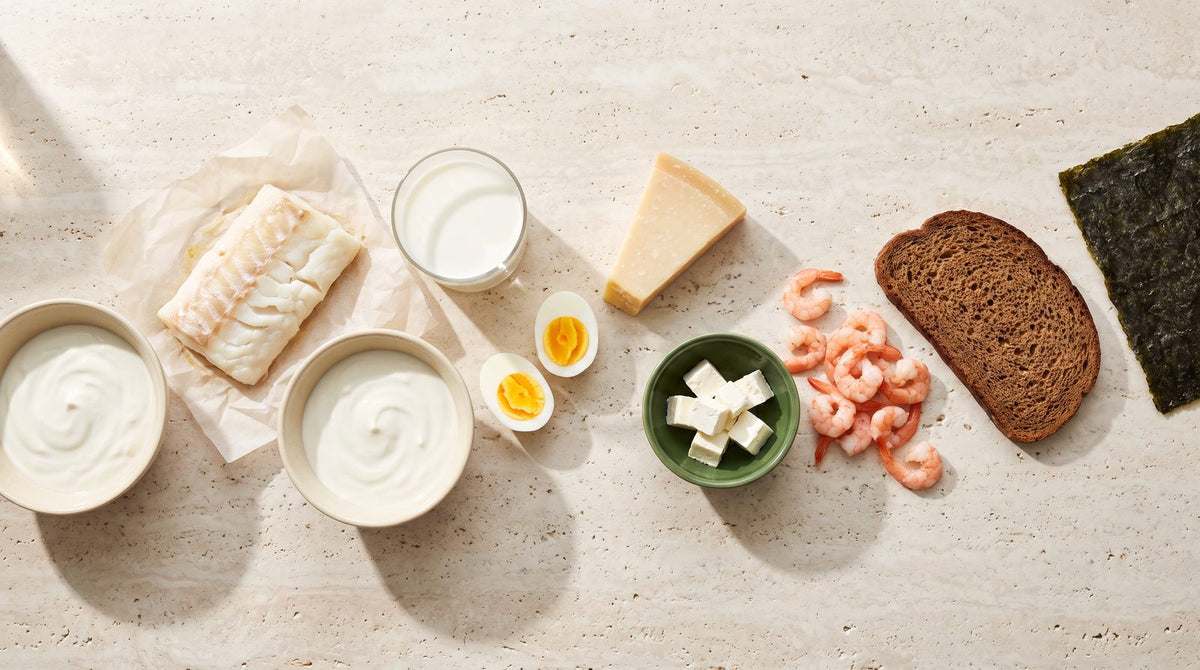 Jodreiche Lebensmittel auf hellem Stein: Kabeljau, Garnelen, Milch, Joghurt, Parmesan, Feta, Eier, Roggenbrot und ein Nori-Blatt