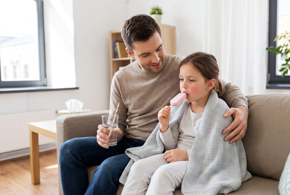 Kind mit Mandelentzündung isst Wassereis auf dem Sofa, der Vater reicht ein Glas Wasser